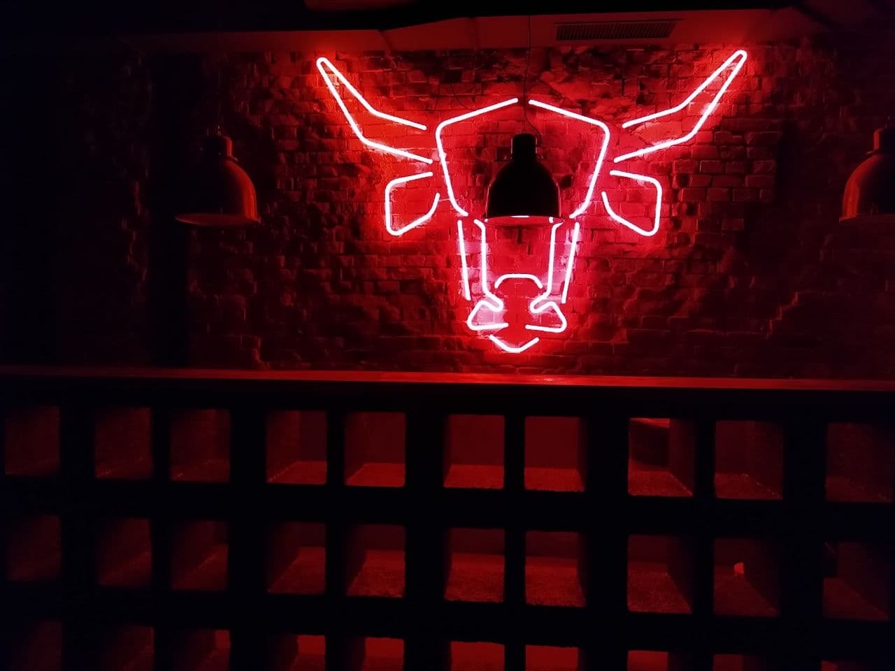 Neonowy wizerunek głowy byka na ceglanej ścianie, oświetlony wiszącymi lampami w ciemnym wnętrzu.