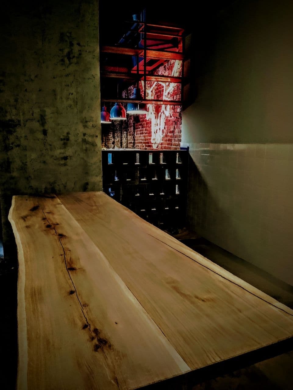 Surowy drewniany blat stołu z naturalnymi krawędziami, ustawiony w odnowionym wnętrzu z odsłoniętą cegłą i betonową ścianą, oświetlony wiszącymi lampami.