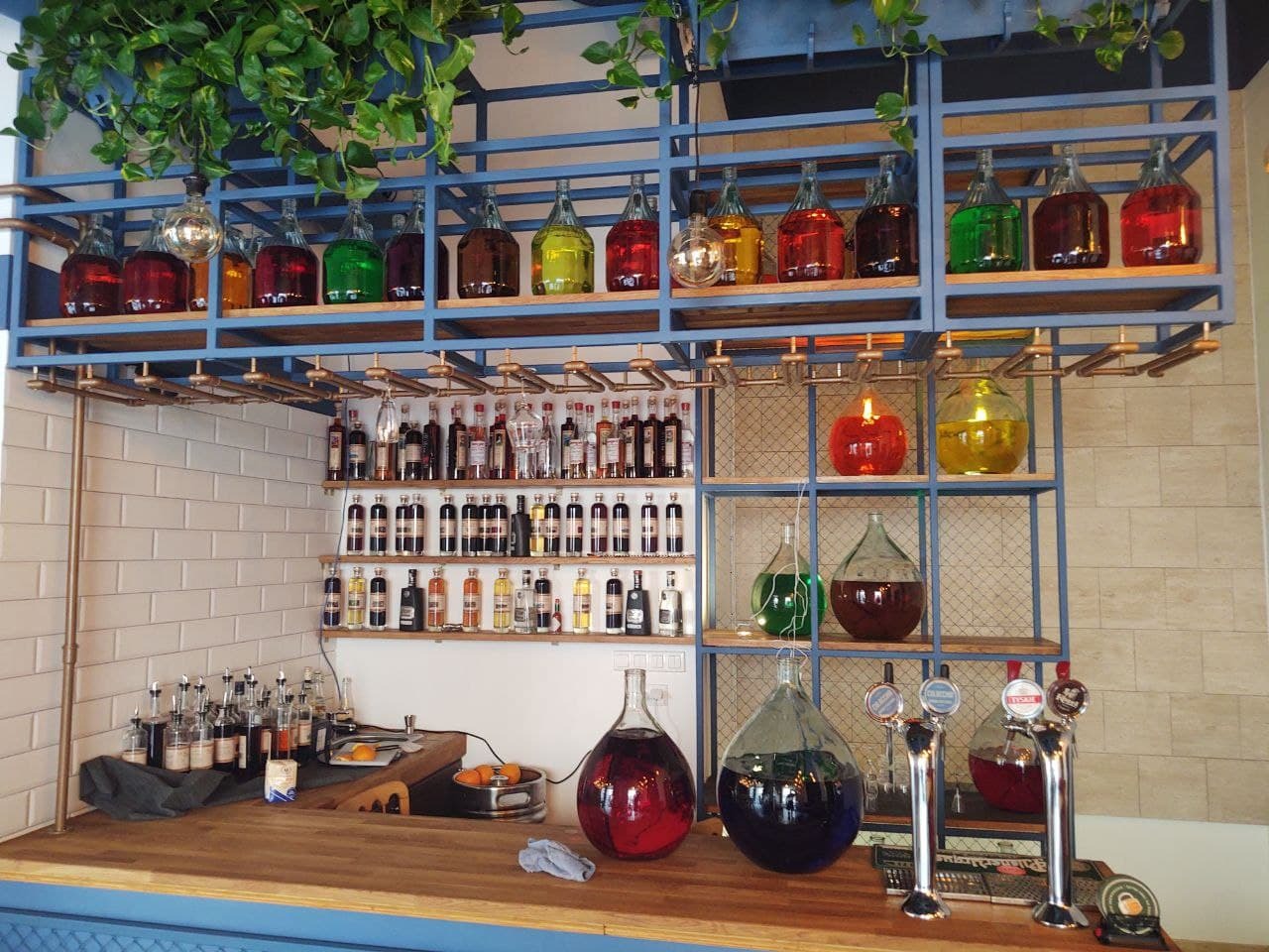 Bar z drewnianym blatem, półkami z kolorowymi butelkami i szklanymi naczyniami, metalowa konstrukcja w kolorze niebieskim, białe płytki na ścianie w tle, rośliny zwisające z góry.