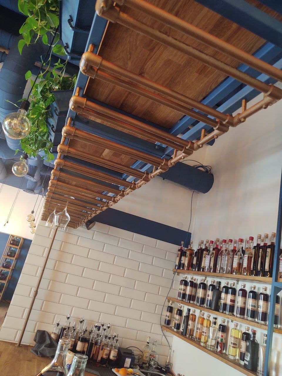 Industrialny wystrój baru: półki z drewnianymi blatami i miedzianymi rurami podwieszone pod sufitem, białe płytki metra na ścianie, butelki alkoholu na półkach, wiszące żarówki i rośliny.