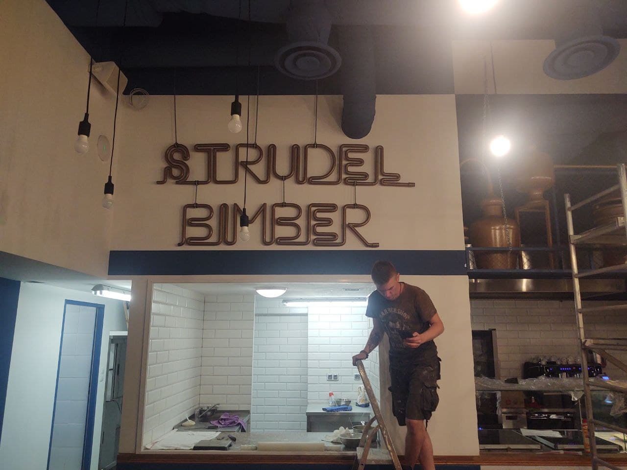 Pracownik na drabinie, prawdopodobnie podczas wykańczania wnętrza lokalu gastronomicznego z białą cegłą, widoczne napisy 'Strudel Bimber' wykonane w formie instalacji z rurek, surowe żarówki...