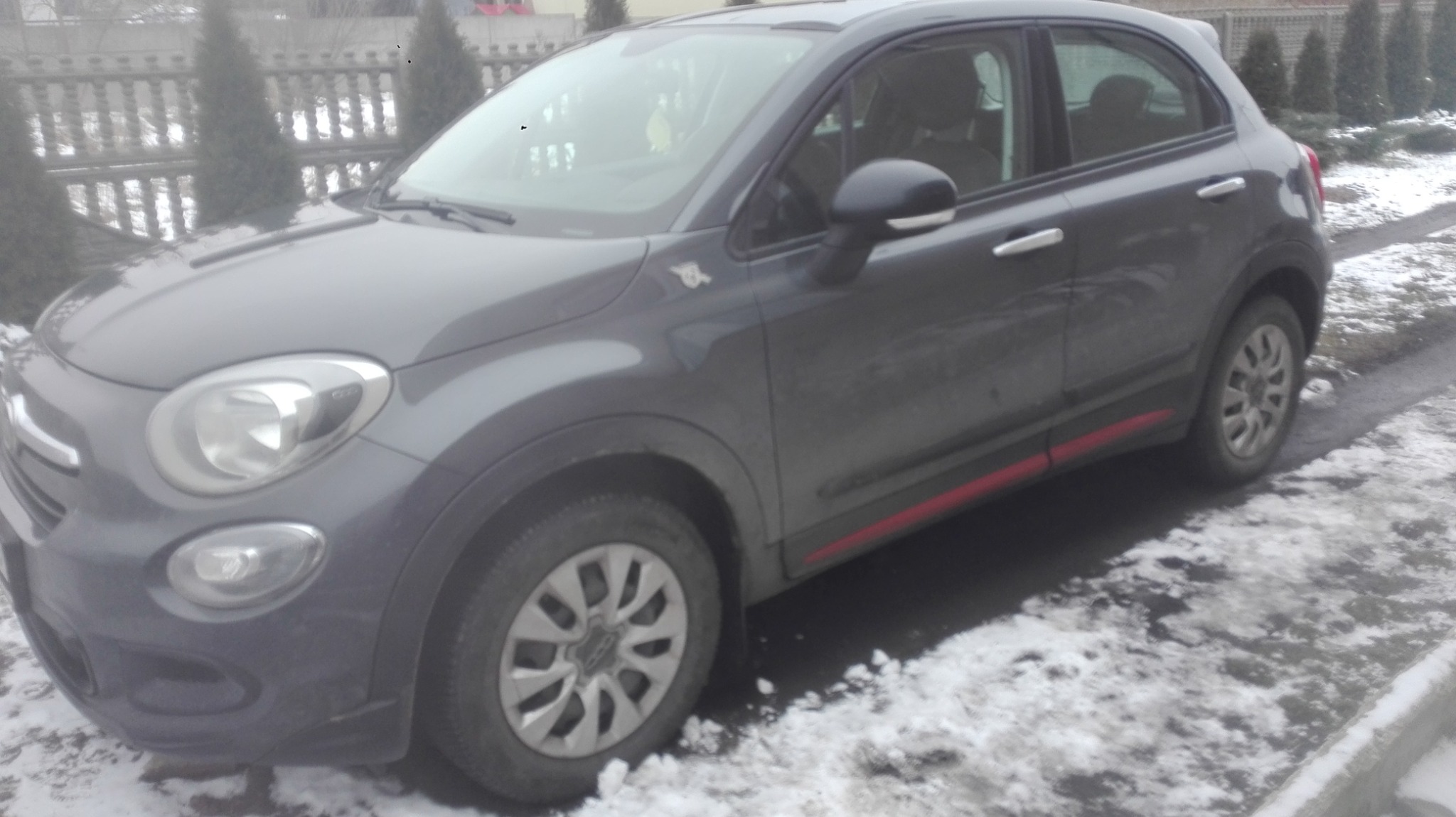 Szary Fiat 500X z czerwoną listwą na progu, stojący na śniegu przed ogrodzeniem z tujami.