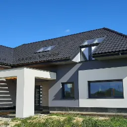 Nowoczesny dom jednorodzinny z grafitową dachówką i elewacją w odcieniach szarości i bieli, widoczne okna dachowe i fragment ogrodu.