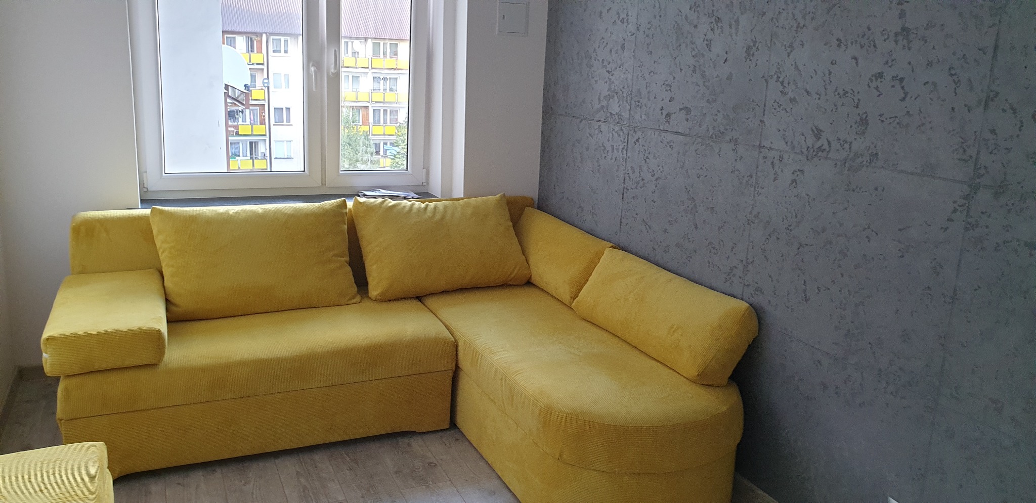 Żółta, narożna sofa z poduszkami na tle szarej ściany z efektem betonu i okna z widokiem na blok.