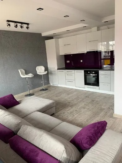 Nowoczesna kuchnia z białymi szafkami, fioletowym panelem szklanym nad blatem, drewnopodobną podłogą i szarą ścianą, uzupełniona beżową sofą z fioletowymi poduszkami i dwoma białymi hokerami.