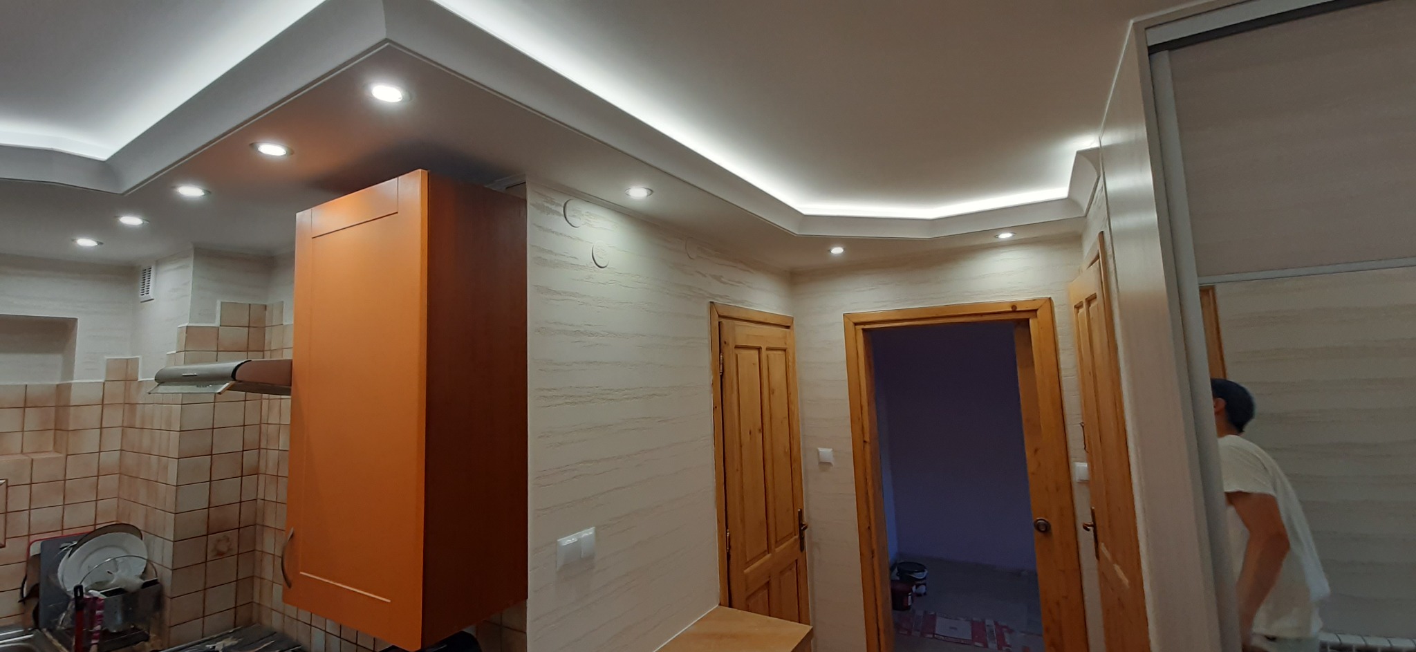 Nowoczesna kuchnia po renowacji z oświetleniem LED w suficie podwieszanym, beżowe ściany z wzorem, brązowe drewniane drzwi i pomarańczowa szafka wisząca.