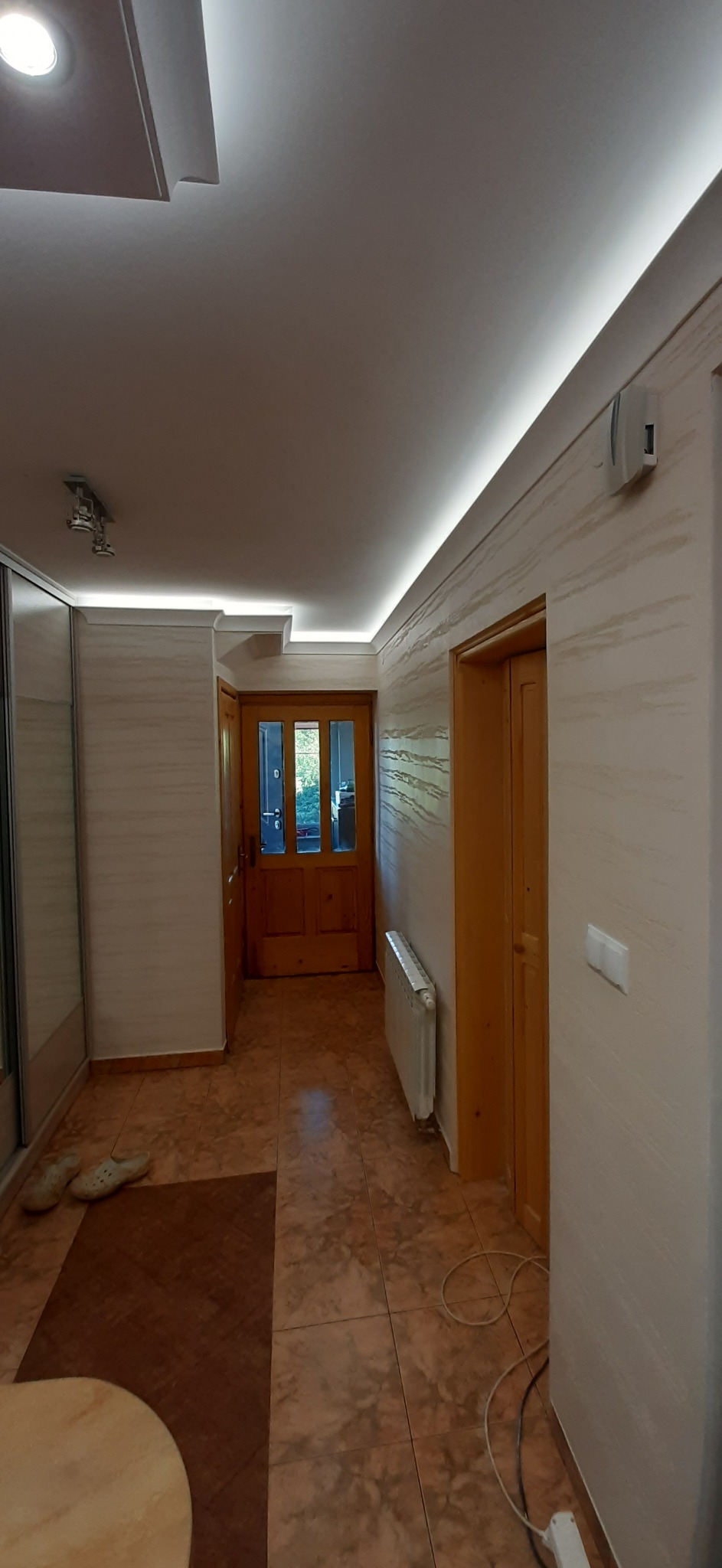 Korytarz z dekoracyjną strukturą ścian w odcieniach beżu, oświetleniem LED podsufitowym, terakotą na podłodze i drewnianymi drzwiami z przeszkleniami.