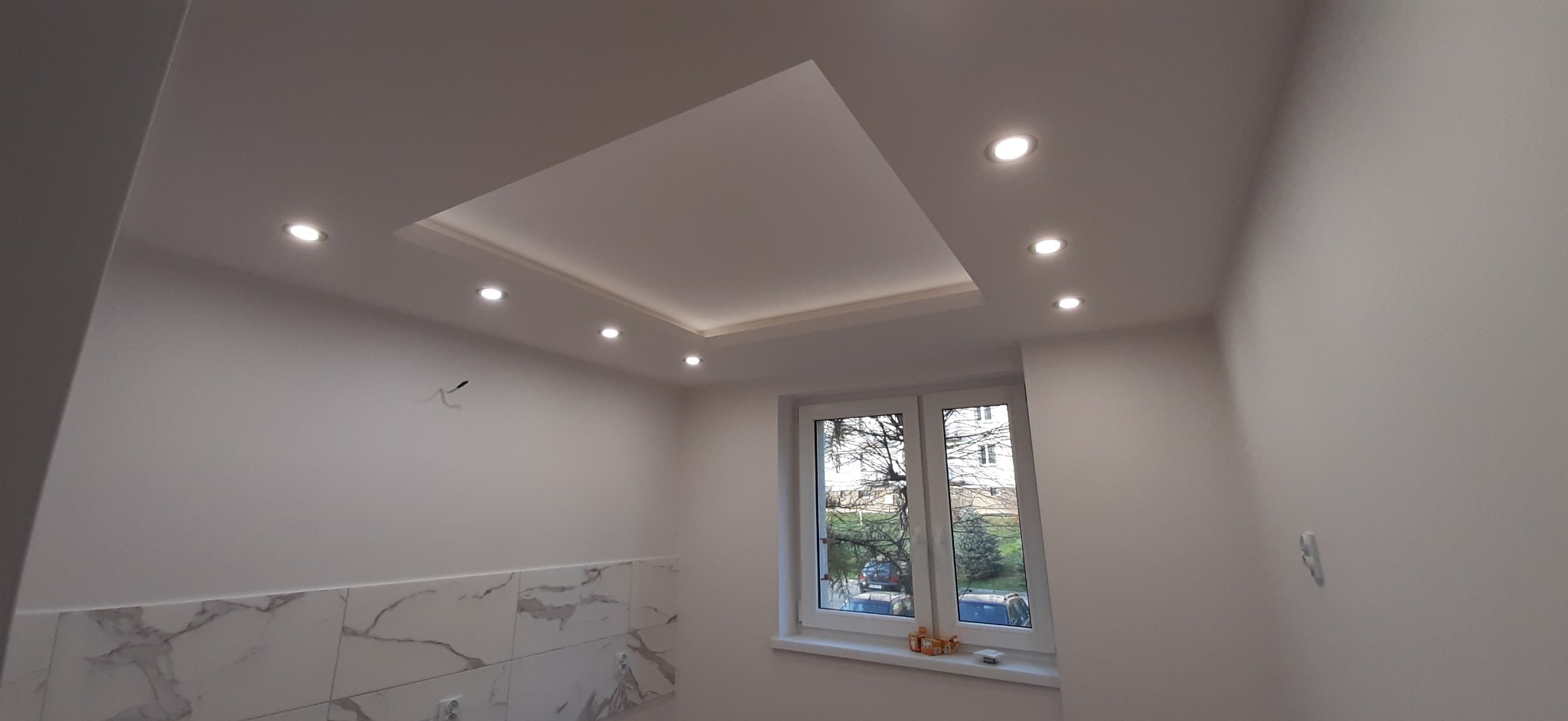 Białe wnętrze po remoncie z podwieszanym sufitem z oświetleniem punktowym i płytkami imitującymi marmur na dole ściany, widok na zewnątrz przez okno.