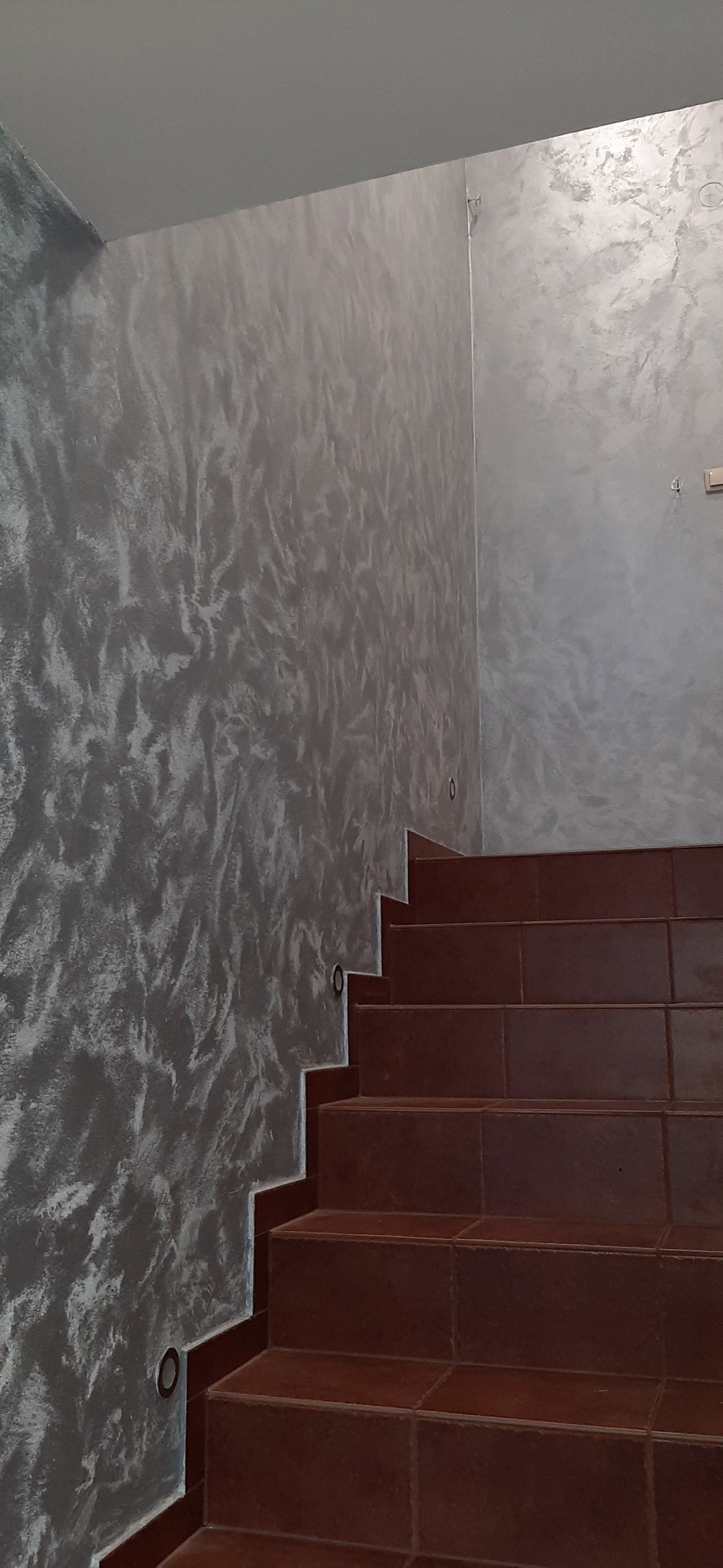 Stylowe schody z brązowych płytek ceramicznych, sąsiadujące ze ścianą pokrytą dekoracyjną szarą farbą o efekcie stiuku weneckiego, z wbudowanymi punktowymi lampkami oświetlającymi bieg schodów.