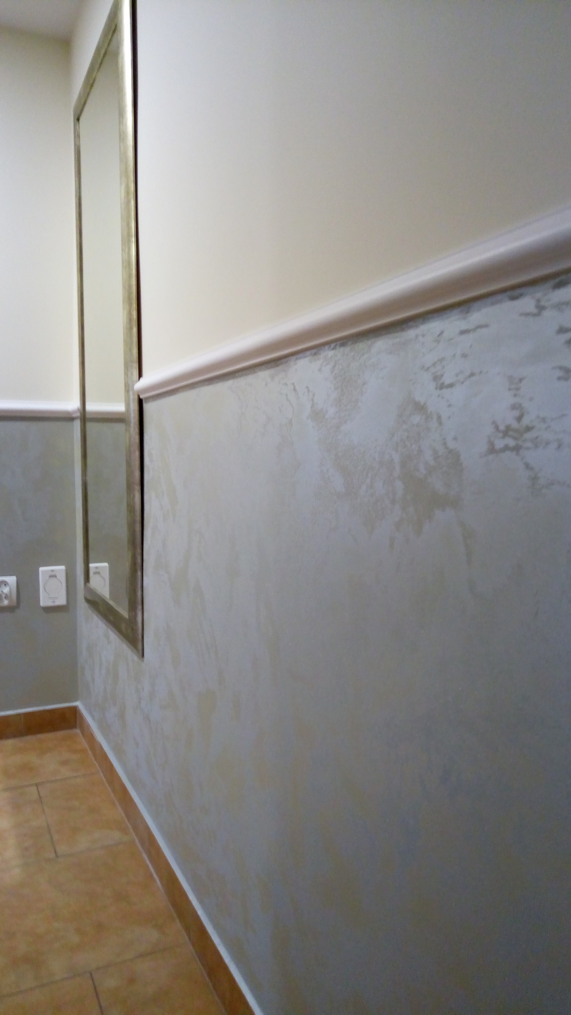 Fragment wykończonego wnętrza łazienki z dekoracyjną ścianą w odcieniach szarości i beżu, listwą ozdobną pod sufitem i lustrem w złotej ramie.