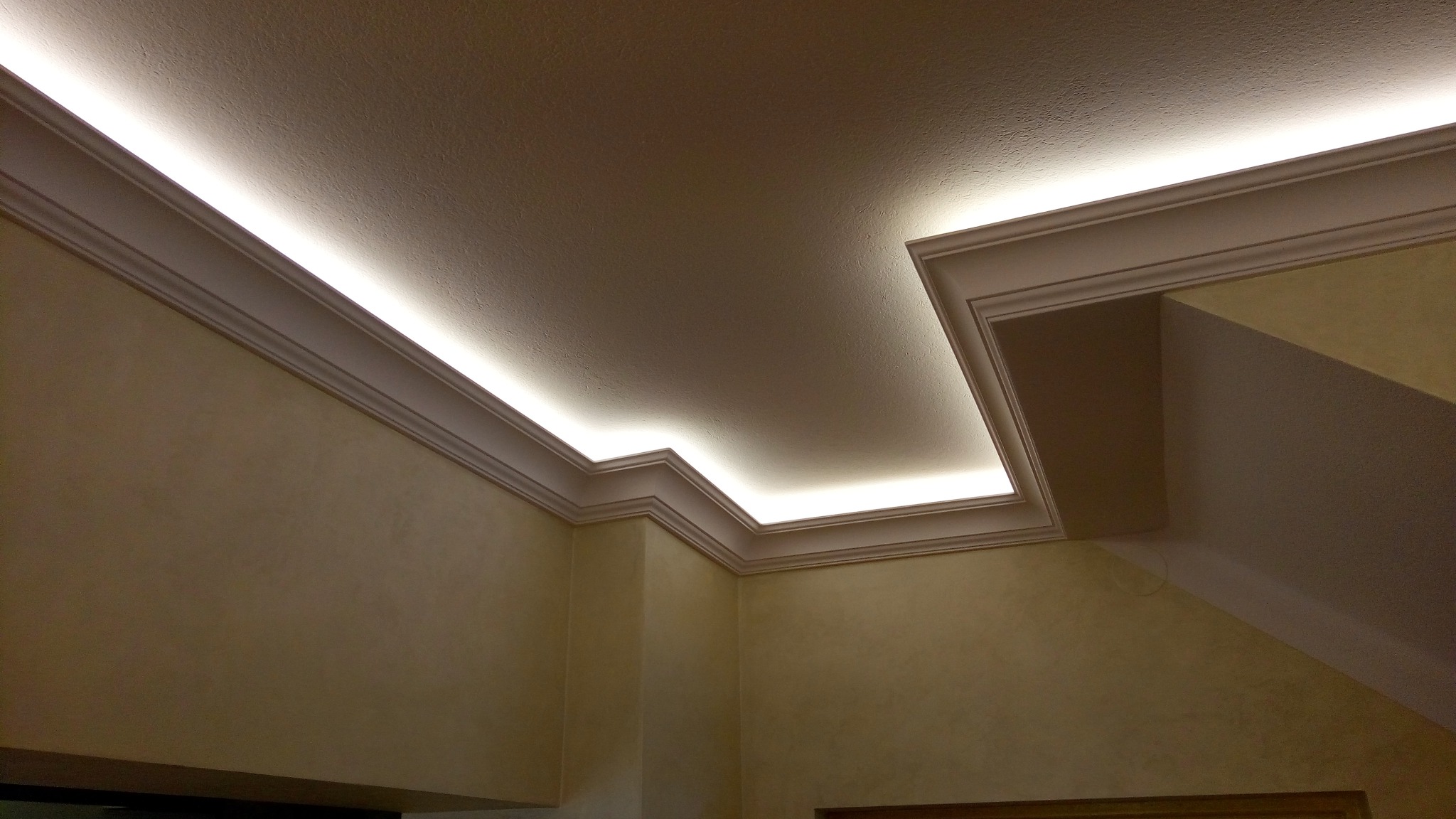 Kąt wewnętrzny pokoju z dekoracyjnym oświetleniem LED ukrytym w listwach przysufitowych, ściany w odcieniach beżu.