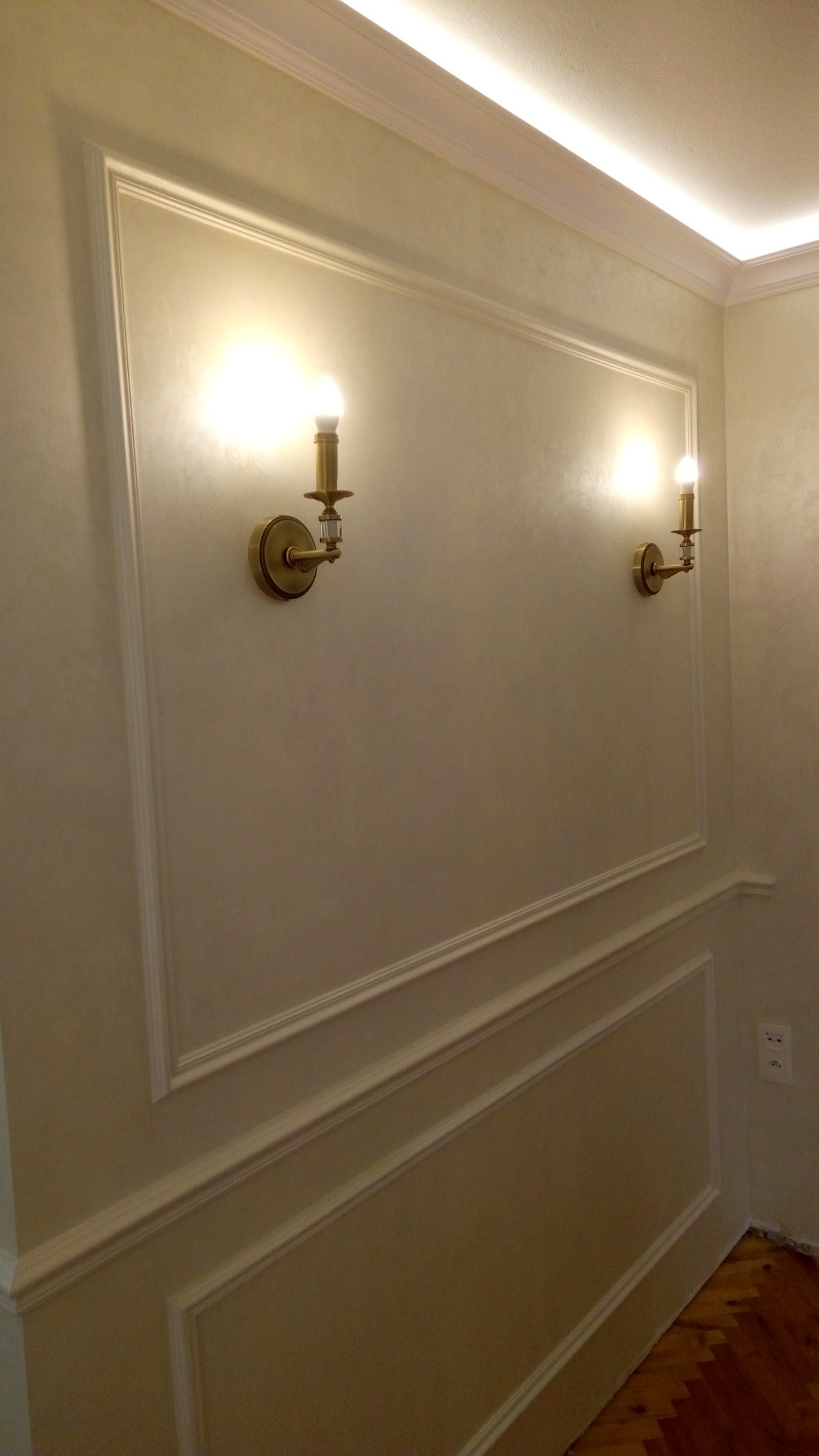 Eleganckie wykończenie ściany z dekoracyjnymi listwami i dwoma złotymi kinkietami z kryształkami, oświetlenie sufitowe LED, parkiet na podłodze.