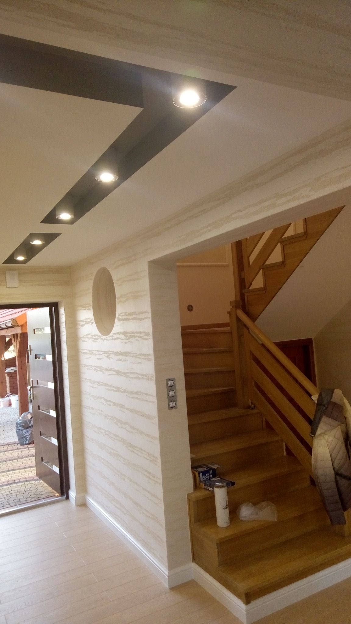 Korytarz z drewnianymi schodami i wzorzystą ścianą z owalnym wgłębieniem, oświetlenie punktowe w suficie, widoczne puszki farb na schodach.