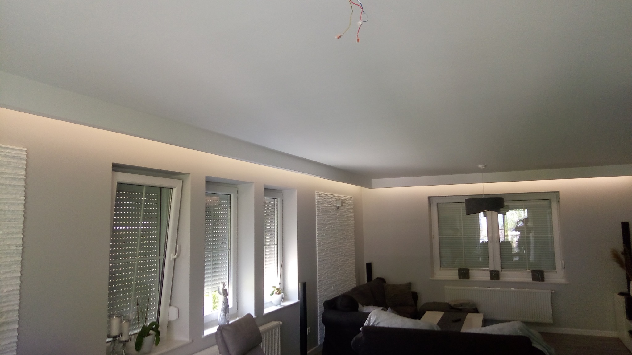 Nowoczesne wnętrze salonu z podświetlanym sufitem, trzema oknami z roletami, dekoracyjną ścianą i częściowo widoczną sofą.