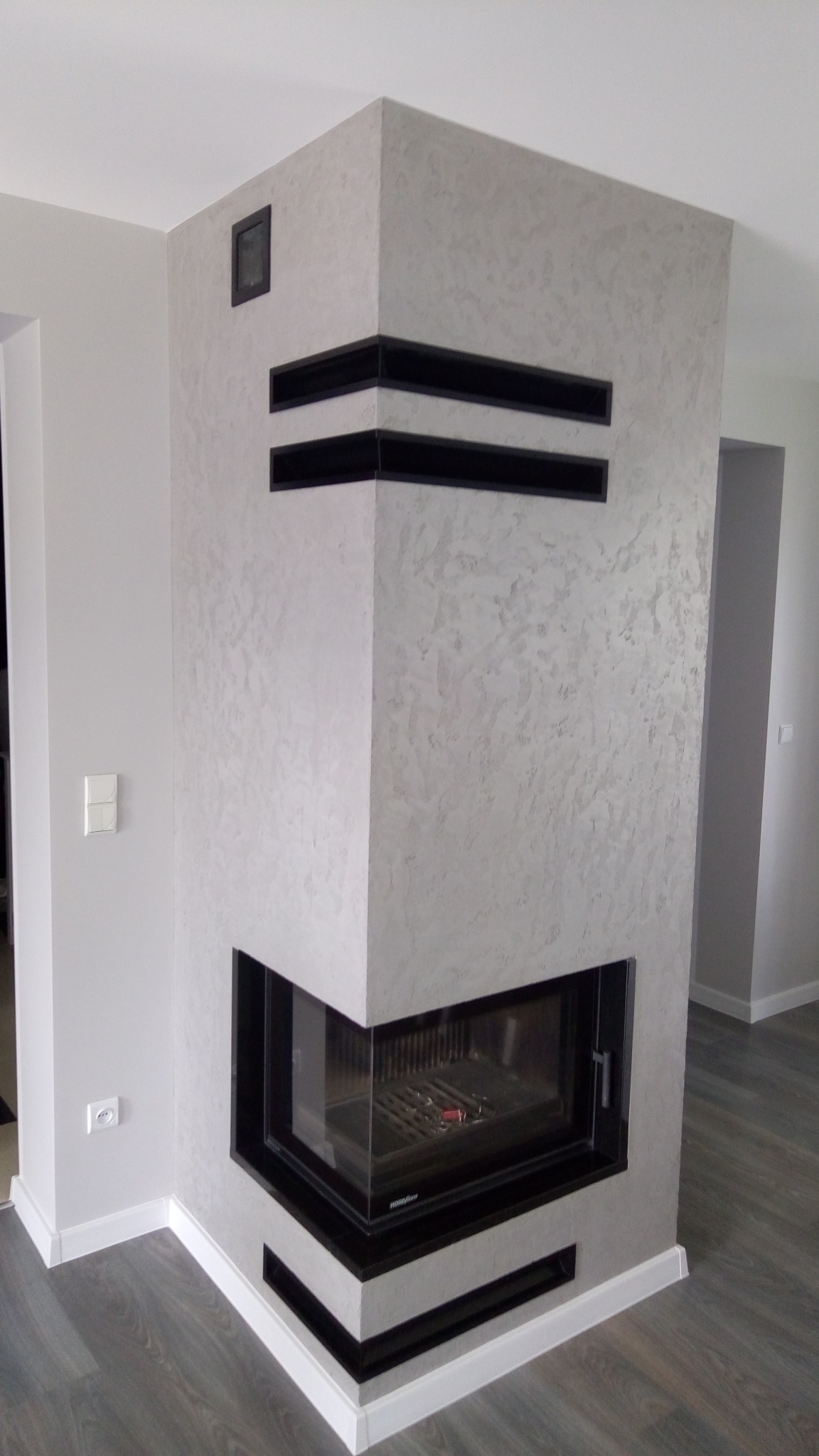 Wykończony narożny kominek z czarnymi akcentami na ścianie o szarej, teksturowanej powierzchni, z widocznymi elementami instalacji elektrycznej.