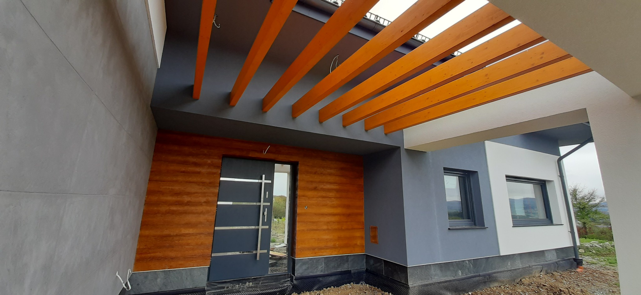 Nowoczesne wejście do domu z drewnianą elewacją i szarą stolarką, widoczna pergola z drewnianych belek.