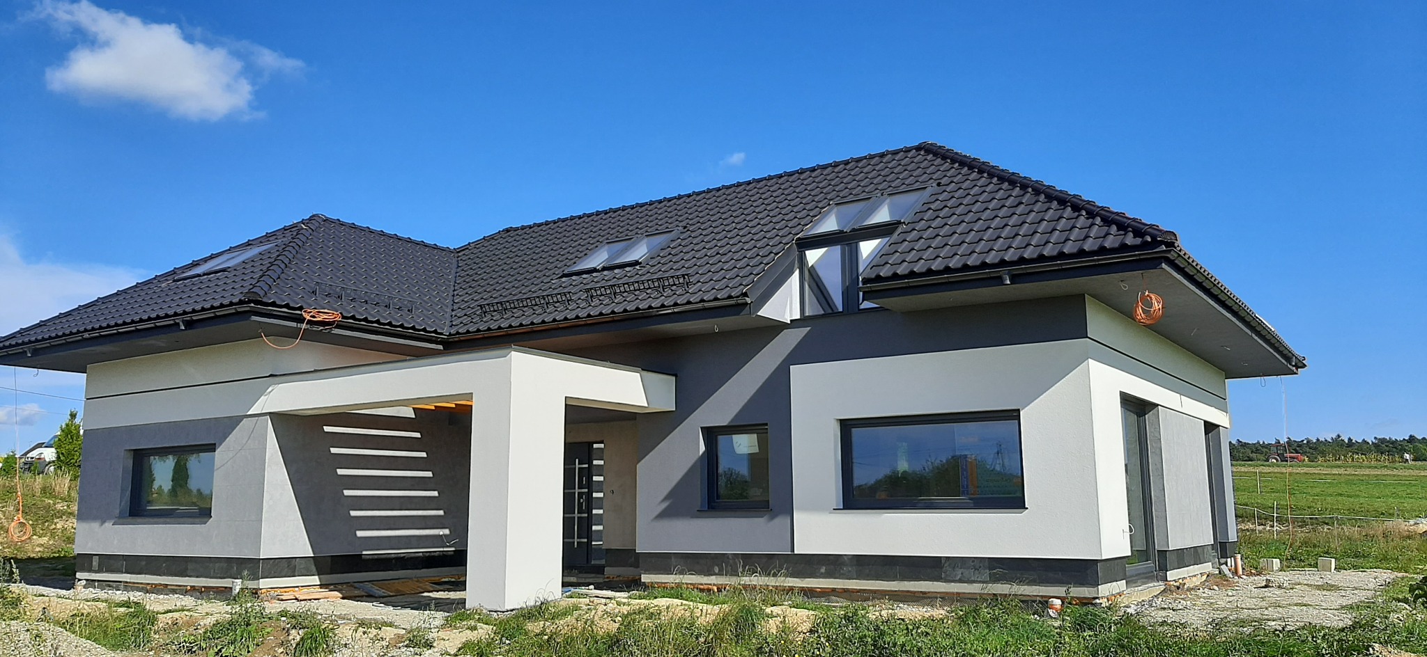 Nowoczesny dom jednorodzinny z grafitową dachówką i elewacją w odcieniach szarości i bieli, widoczne okna dachowe i fragment ogrodu.