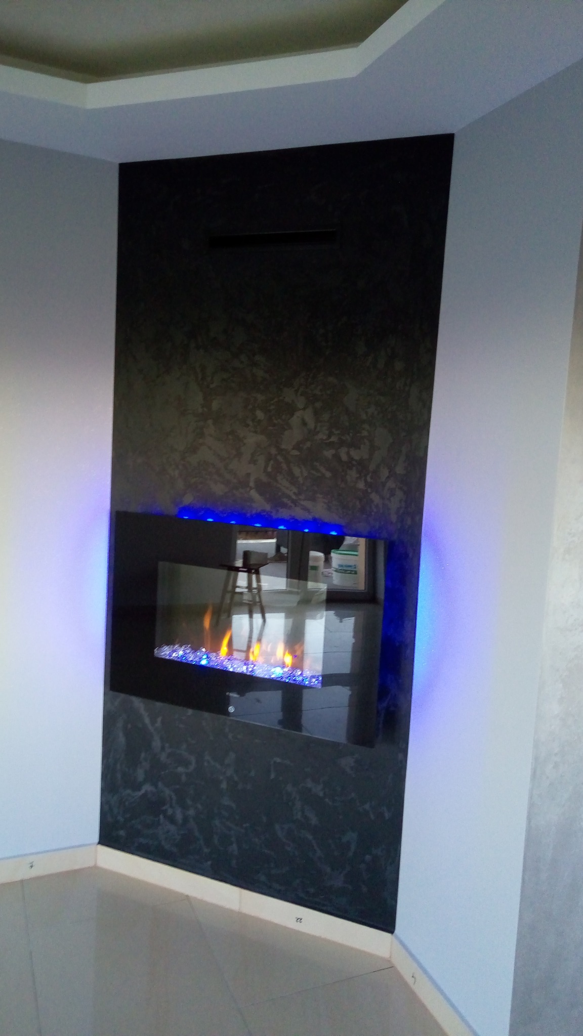 Ściana z dekoracyjną, ciemną okładziną, w której wbudowany jest elektryczny kominek z niebieskim podświetleniem LED i imitacją płomieni, odbicie krzesła w szkle kominka.