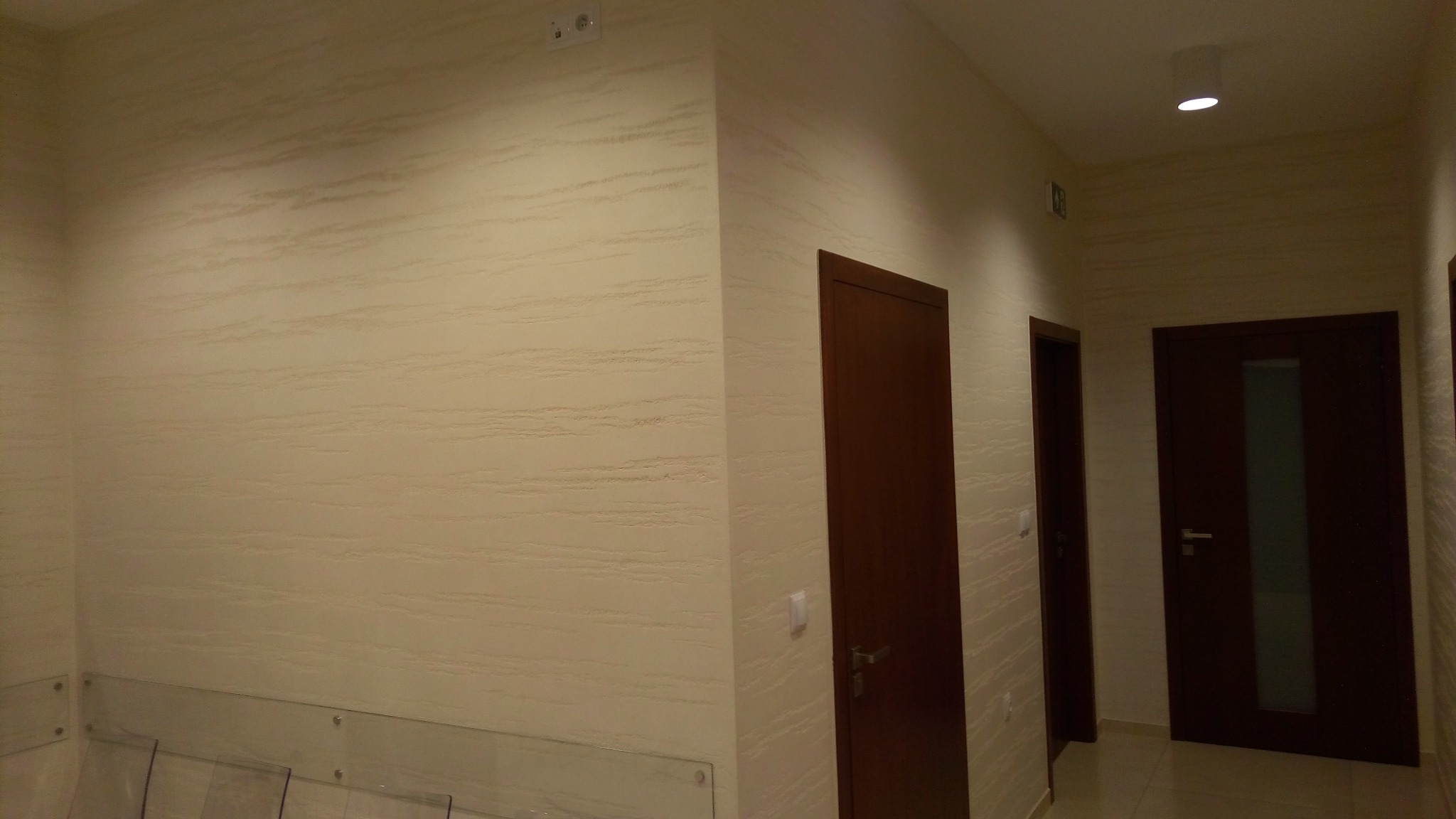 Korytarz z kremowymi ścianami o dekoracyjnym, poziomym wzorze, z trzema ciemnymi, drewnianymi drzwiami i szklaną balustradą w dolnej części kadru.