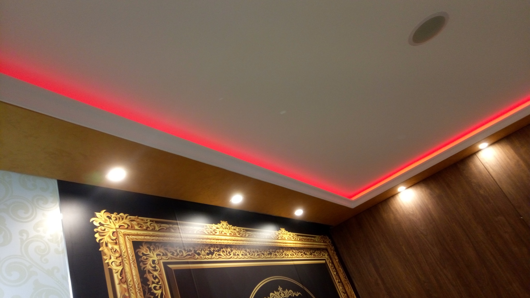 Sufit podwieszany z oświetleniem LED w kolorze czerwonym, widoczne ozdobne wykończenia ścian i sufitu, oraz fragment dekoracyjnego obrazu w złotej ramie.