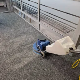 Szorowarka do dywanów w trakcie pracy na szarym dywanie w biurze, widoczny fragment umytej powierzchni oraz przeszklenia biurowe.