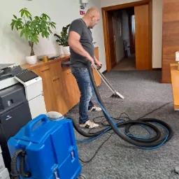 Mężczyzna czyści wykładzinę w biurze za pomocą profesjonalnego odkurzacza piorącego z niebieskim zbiornikiem, widoczne biurko, drukarka i rośliny doniczkowe w tle.