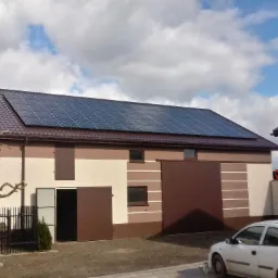 Panele słoneczne zamontowane na dachu budynku gospodarczego w Turku, widoczne z poziomu gruntu, z przodu zaparkowany biały samochód osobowy.