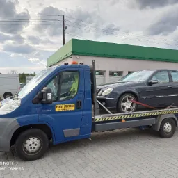 Niebieska laweta z czarnym Mercedesem na platformie, zabezpieczonym czerwonym pasem, na tle budynku z zielonym dachem i napisem 'SKLEP', na parkingu z kostki brukowej.