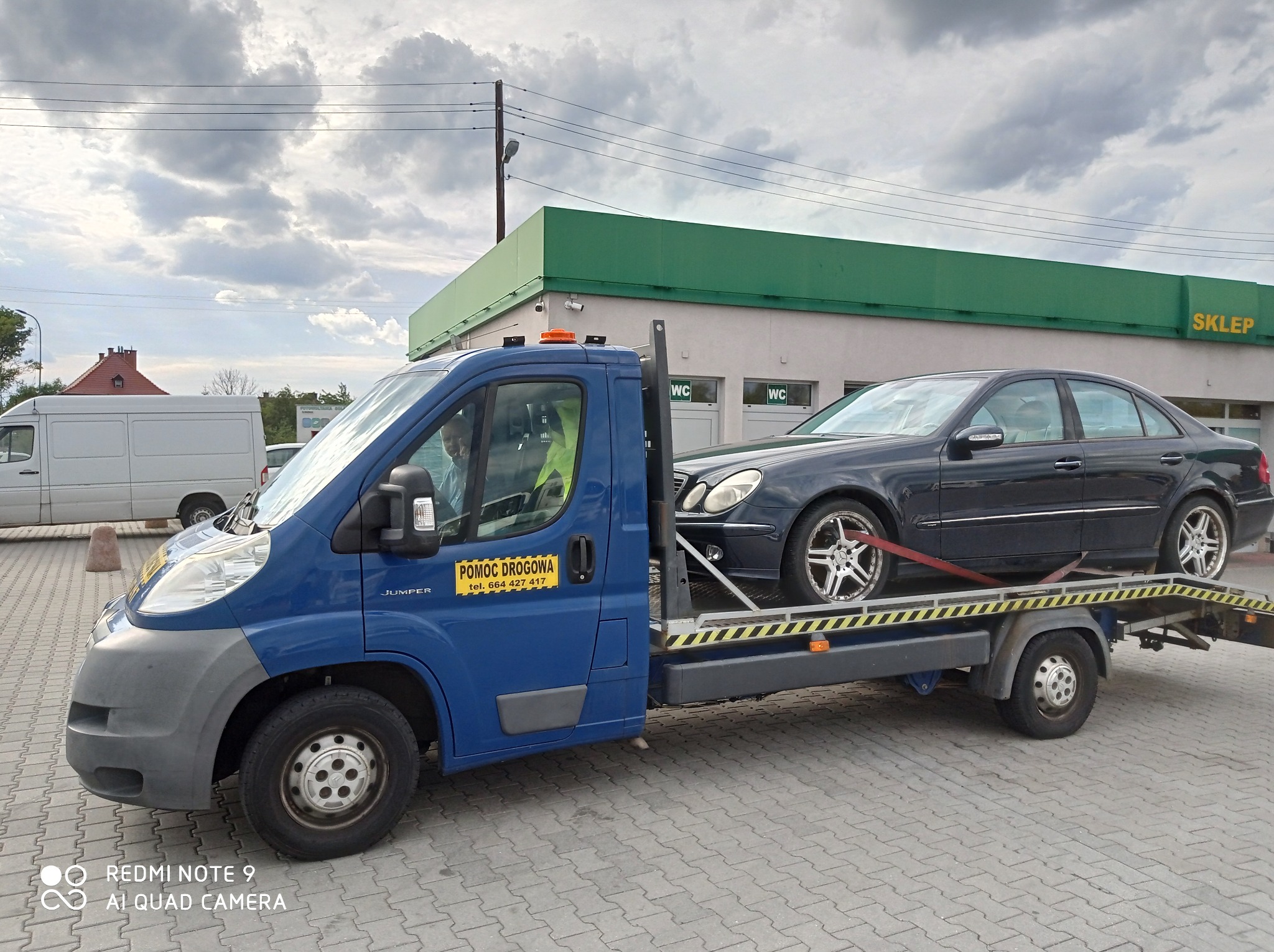 Niebieska laweta z czarnym Mercedesem na platformie, zabezpieczonym czerwonym pasem, na tle budynku z zielonym dachem i napisem 'SKLEP', na parkingu z kostki brukowej.