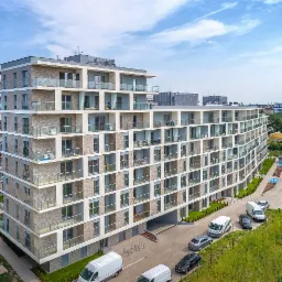 Wielopiętrowy budynek mieszkalny z licznymi balkonami i jasnymi ramami okiennymi, widok z góry na parking z samochodami dostawczymi.