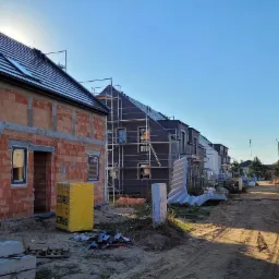 Nowe domy w trakcie budowy: jeden z czerwoną cegłą, drugi z szarą elewacją i rusztowaniem, w tle silosy na materiały budowlane.