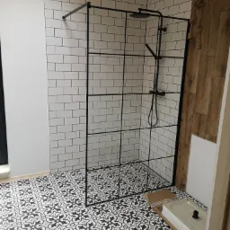 Nowoczesna kabina prysznicowa z czarnymi profilami na tle białych płytek typu metro i wzorzystej, czarno-białej podłogi. Część ściany wykończona drewnianymi panelami, widoczny fragment umywalki...