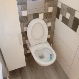 Biała toaleta z otwartą klapą w nowoczesnej łazience z geometrycznymi płytkami na ścianie i szarą podłogą imitującą drewno.