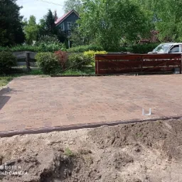 Ułożona kostka brukowa w odcieniach brązu, z widocznym obrzeżem i przygotowanym podłożem z ziemi, w tle brama wjazdowa i zaparkowany samochód dostawczy.