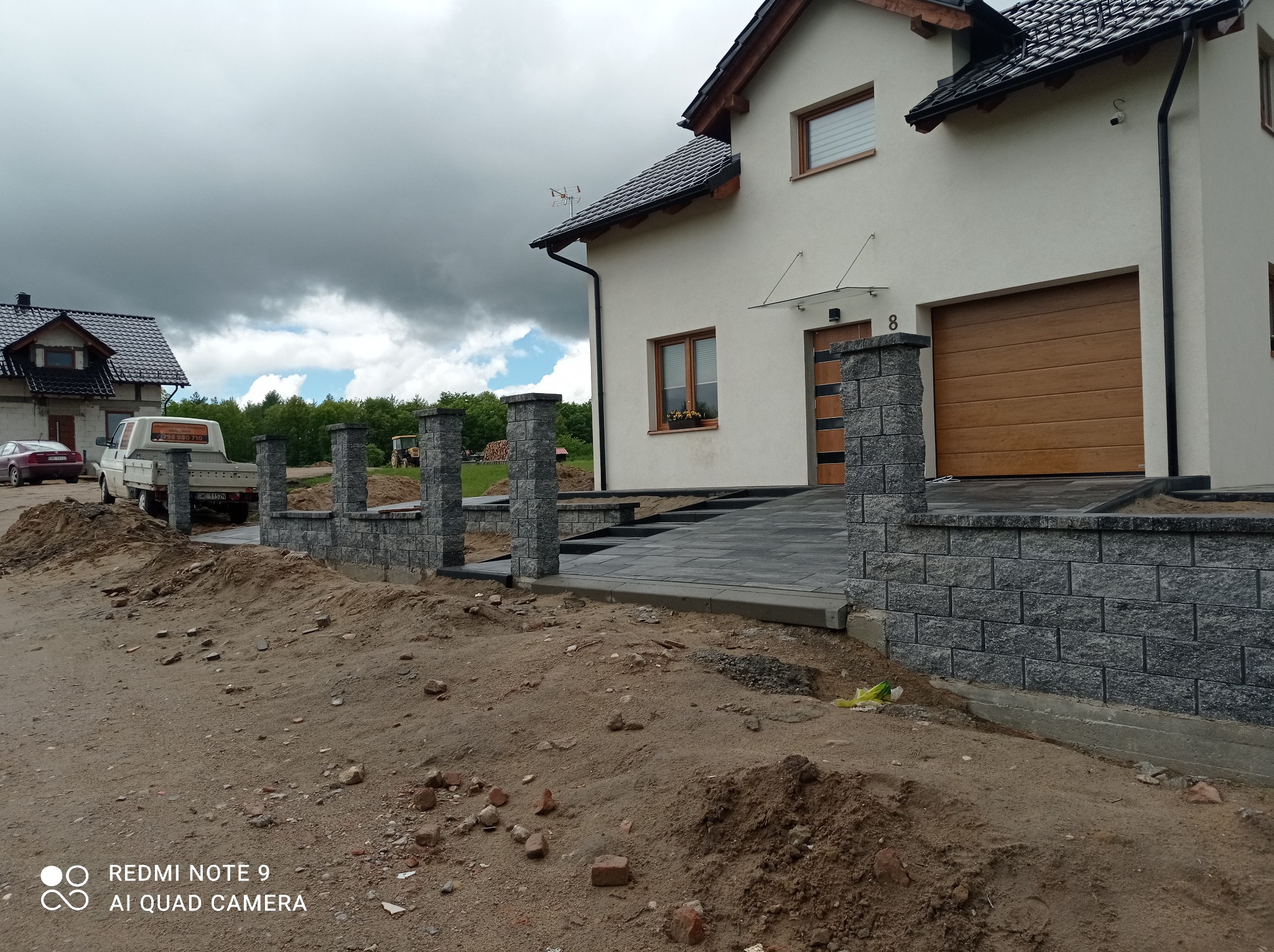 Nowoczesny dom z szarej kostki brukowej prowadzącej do drzwi i bramy garażowej, z niskim ogrodzeniem z kamiennych słupków i murków, widoczny samochód dostawczy i traktor w tle, pochmurne niebo.