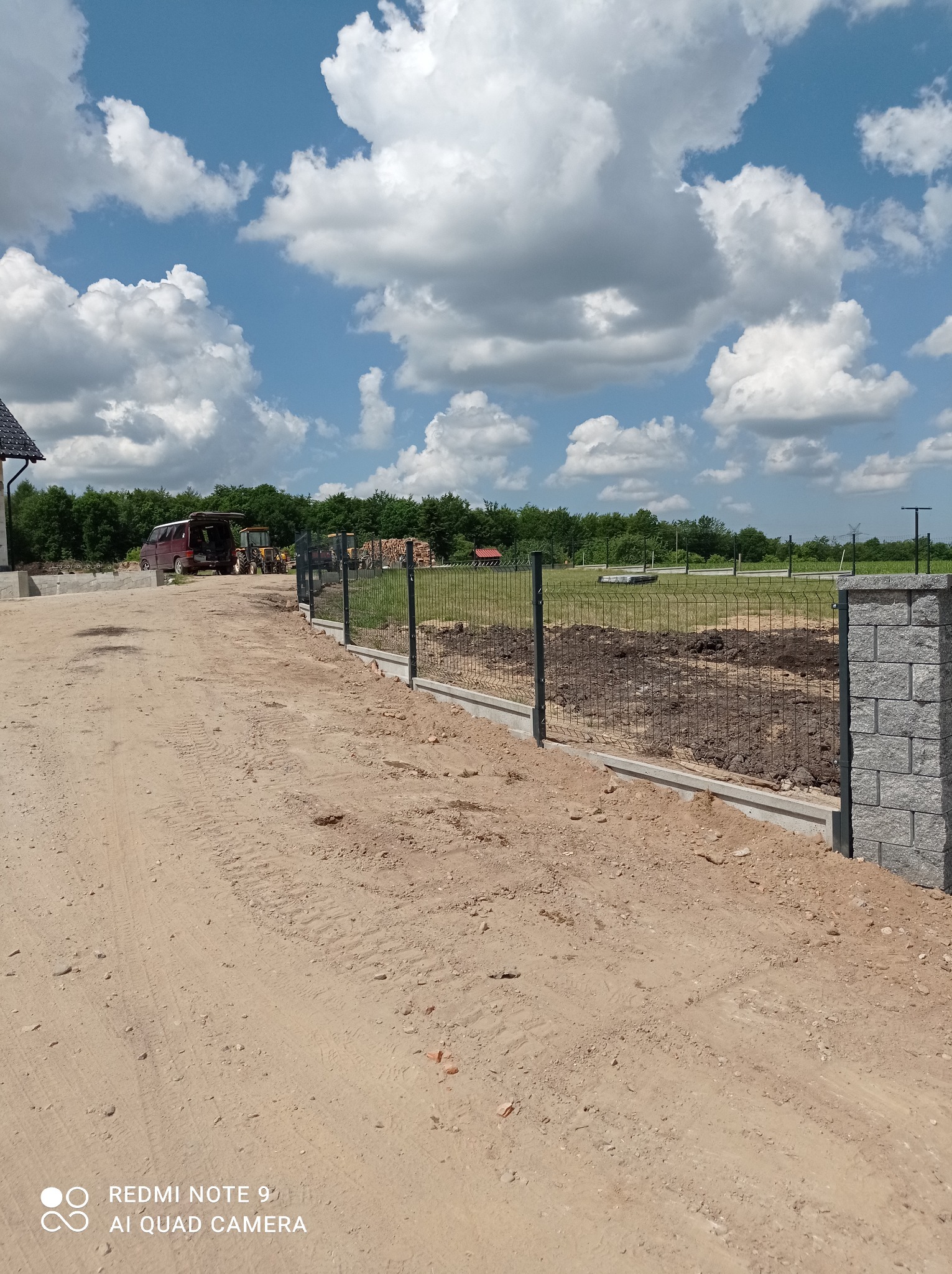 Wznoszone ogrodzenie panelowe z podmurówką betonową i słupkami z imitacji kamienia, widoczne maszyny budowlane w tle, dzień słoneczny.