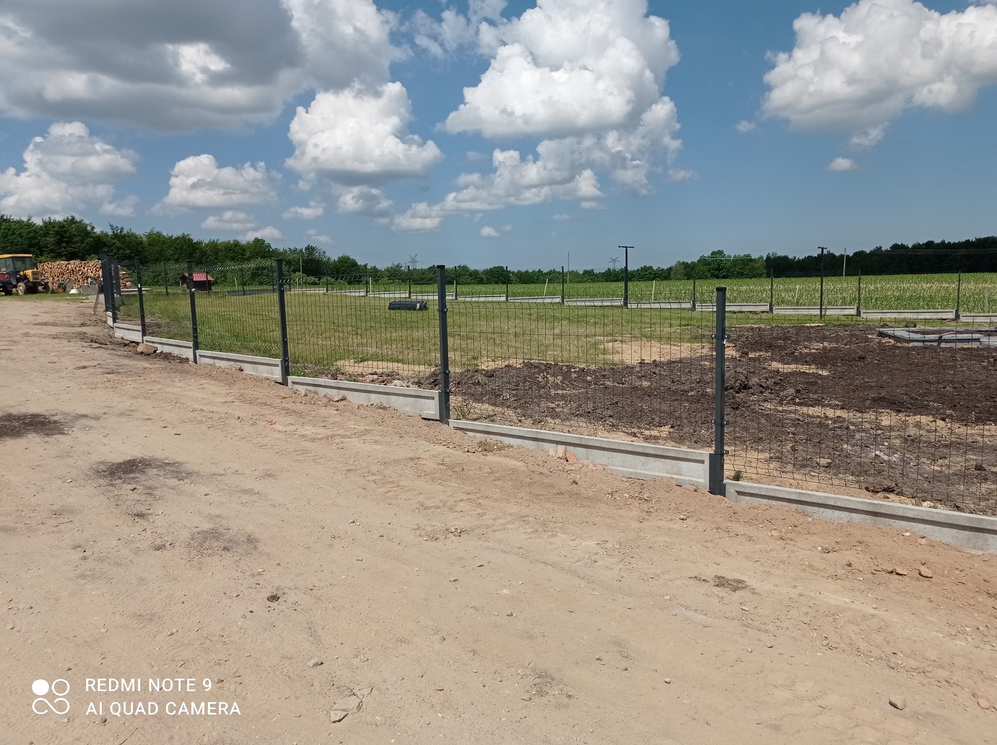 W trakcie budowy ogrodzenia panelowego z podmurówką betonową na działce, widoczne słupki i siatka, ziemia przygotowana pod dalsze prace, w tle pole uprawne i las.