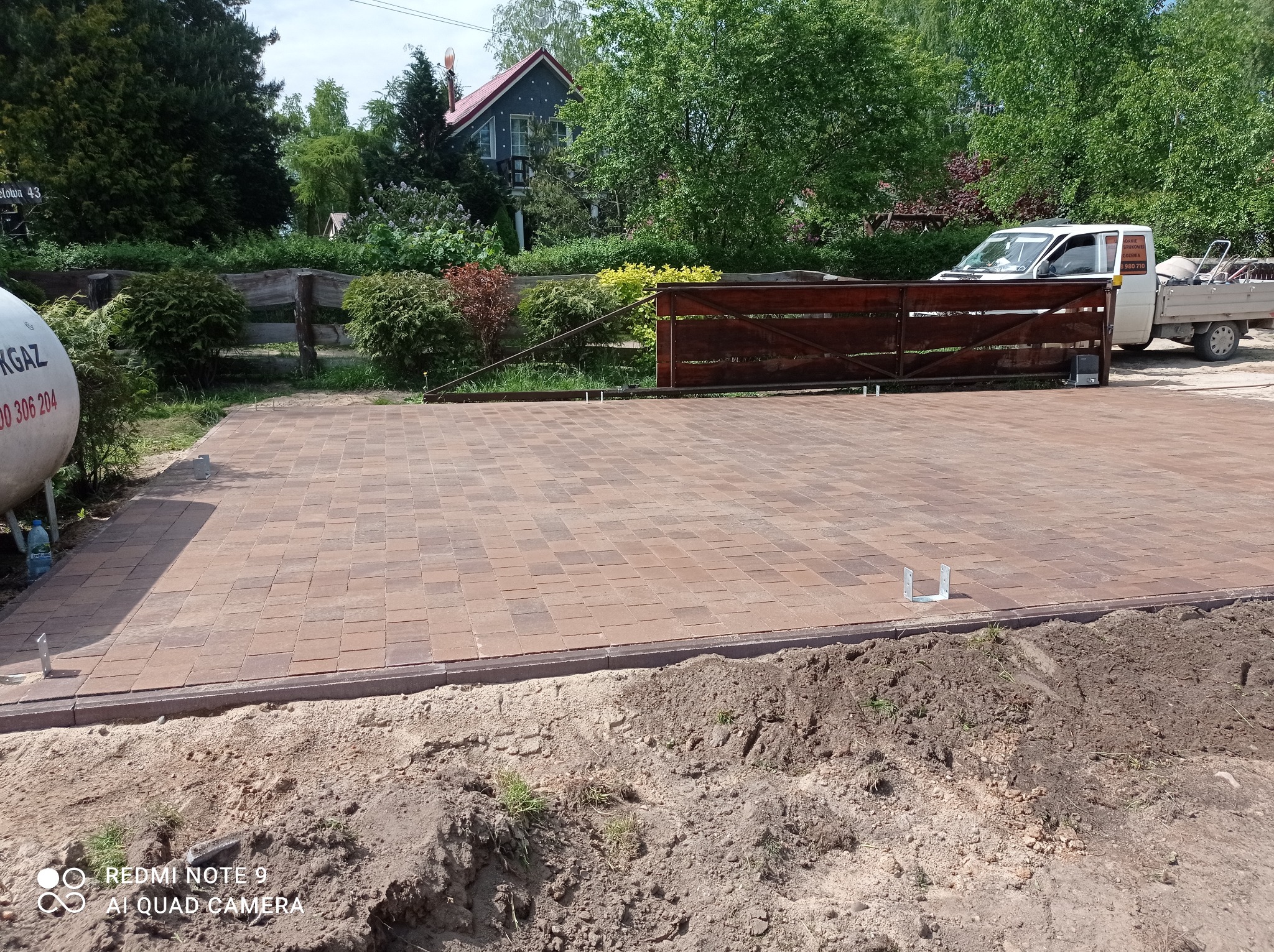 Ułożona kostka brukowa w odcieniach brązu, z widocznym obrzeżem i przygotowanym podłożem z ziemi, w tle brama wjazdowa i zaparkowany samochód dostawczy.