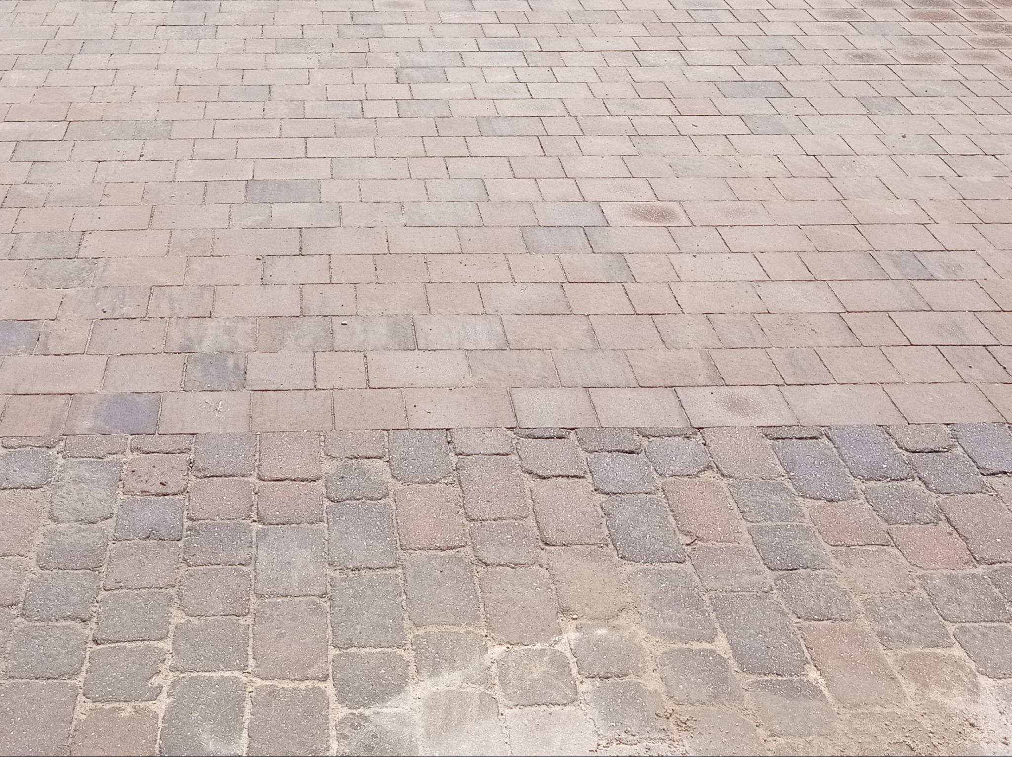 Ułożona nawierzchnia z kostki brukowej w dwóch odcieniach: jaśniejszym beżowym w górnej części i ciemniejszym szarym w dolnej, z widocznymi spoinami wypełnionymi piaskiem.