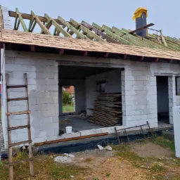 Niedokończony budynek z bloczków betonowych z częściowo ukończonym drewnianym dachem i kominem zabezpieczonym żółtą folią, widoczna drabina oparta o ścianę i paleta bloczków Solbet.