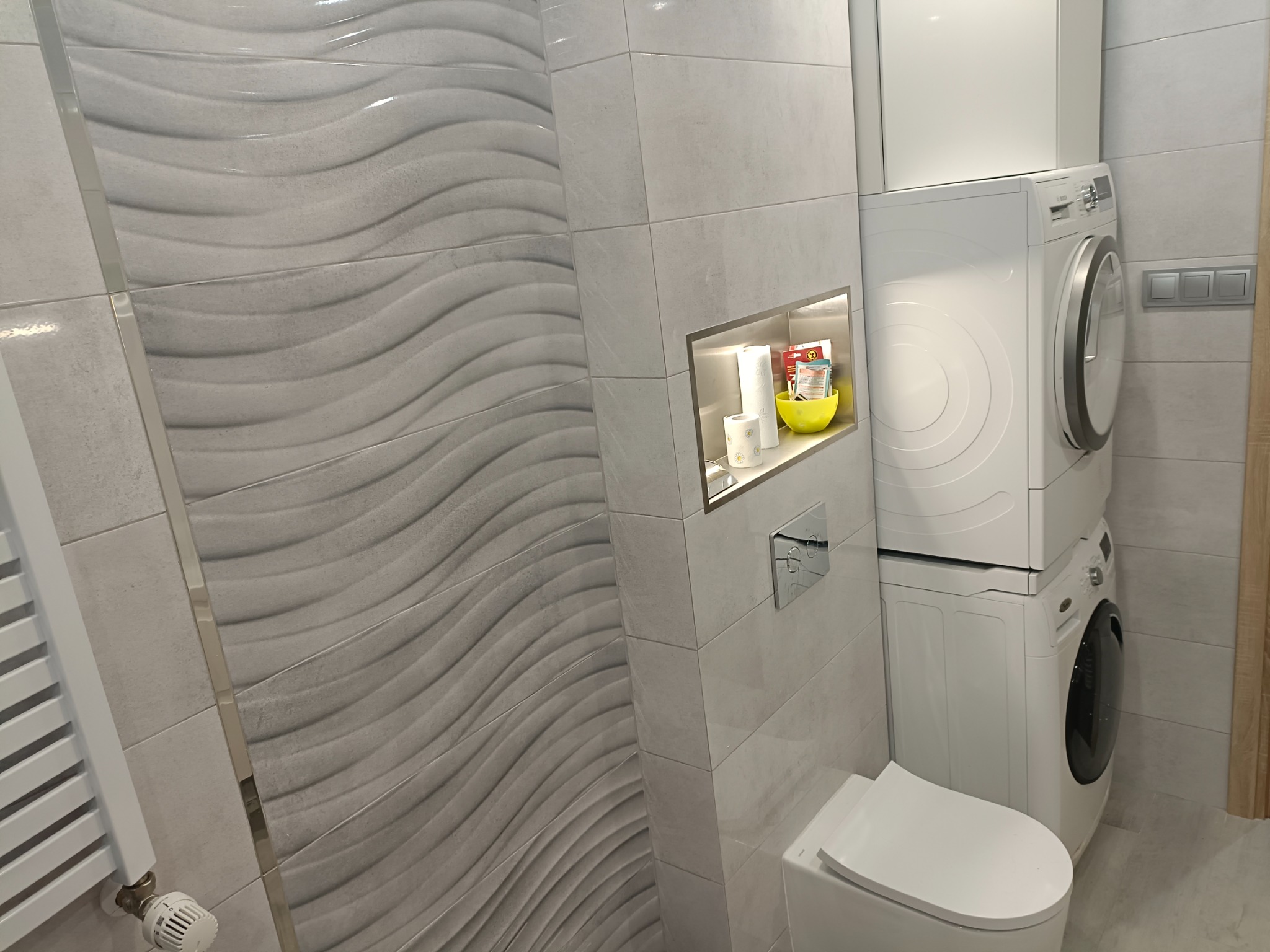 Szara łazienka z falistymi płytkami, wnęką z akcesoriami, toaletą i pralko-suszarką ustawioną w pionie.