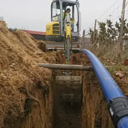 Żółta mini koparka Wacker Neuson umieszczona nad wykopem, w tle pryzma ziemi i budynek gospodarczy, w wykopie widoczny fragment niebieskiej rury.