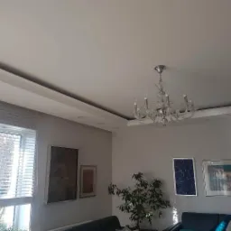 Jasne wnętrze salonu z gładkimi, szarymi ścianami, białym sufitem z listwami i kryształowym żyrandolem, oświetlonymi przez okno z żaluzjami.