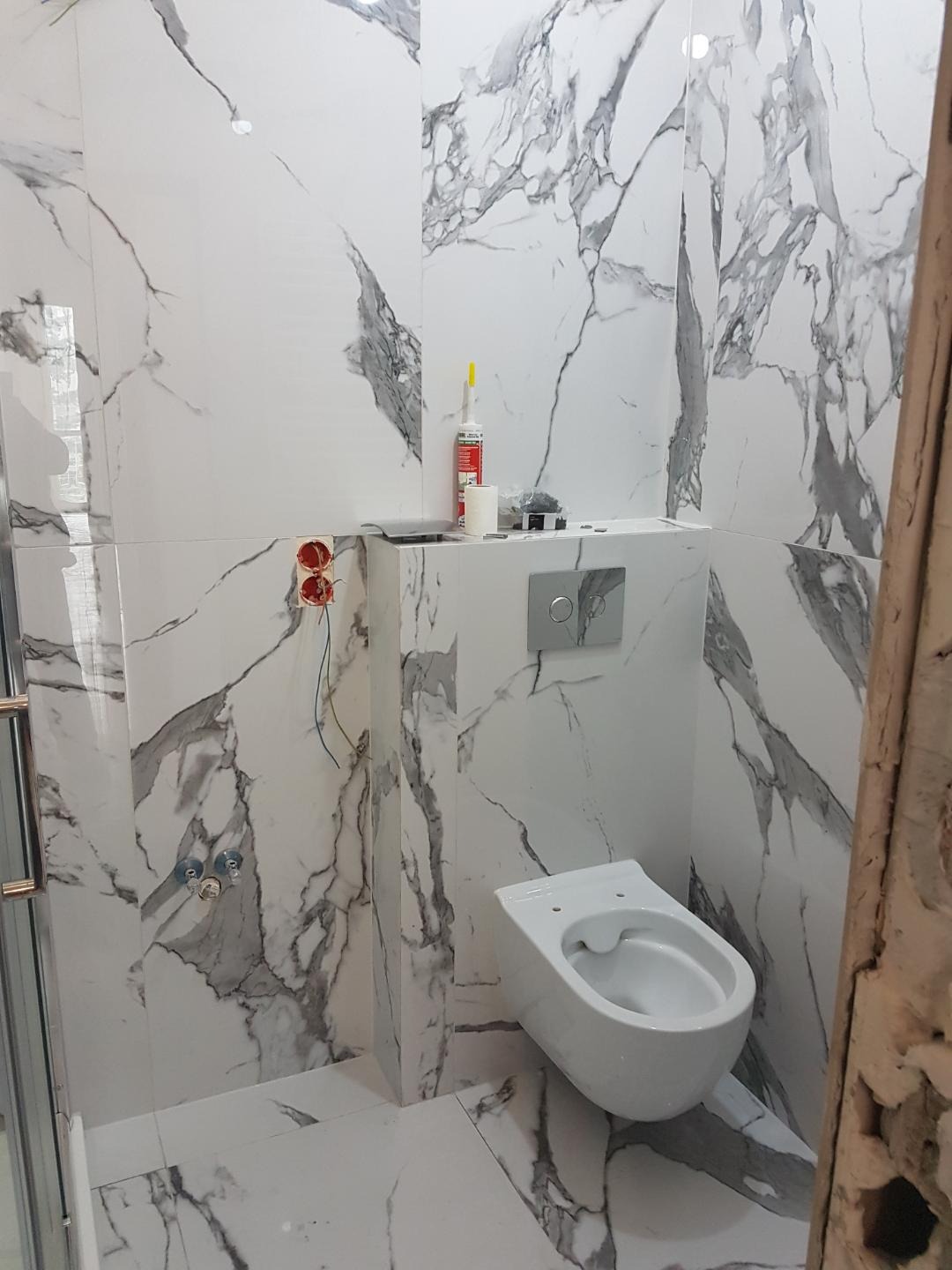 Wnętrze łazienki w trakcie montażu podwieszanej toalety, z widocznymi płytkami imitującymi marmur, puszkami elektrycznymi i akcesoriami montażowymi.