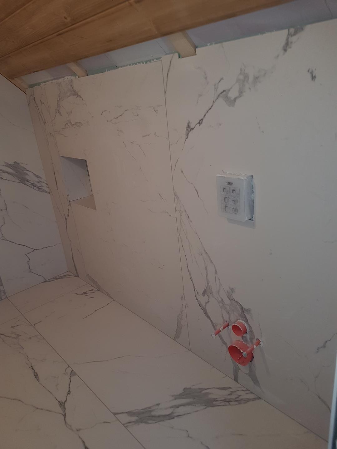 Łazienka w trakcie wykańczania: białe płytki imitujące marmur na ścianach i podłodze, widoczne przyłącza wodne i wnęka w ścianie, surowy drewniany strop.