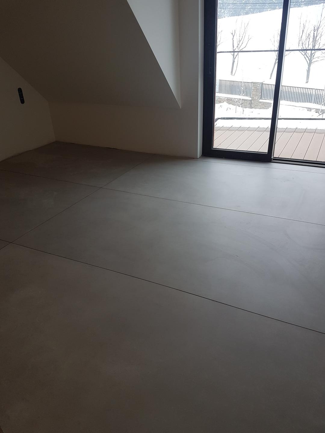 Szare, wykończone podłoże w pokoju na poddaszu, widok na zimowy krajobraz przez przesuwne drzwi balkonowe.