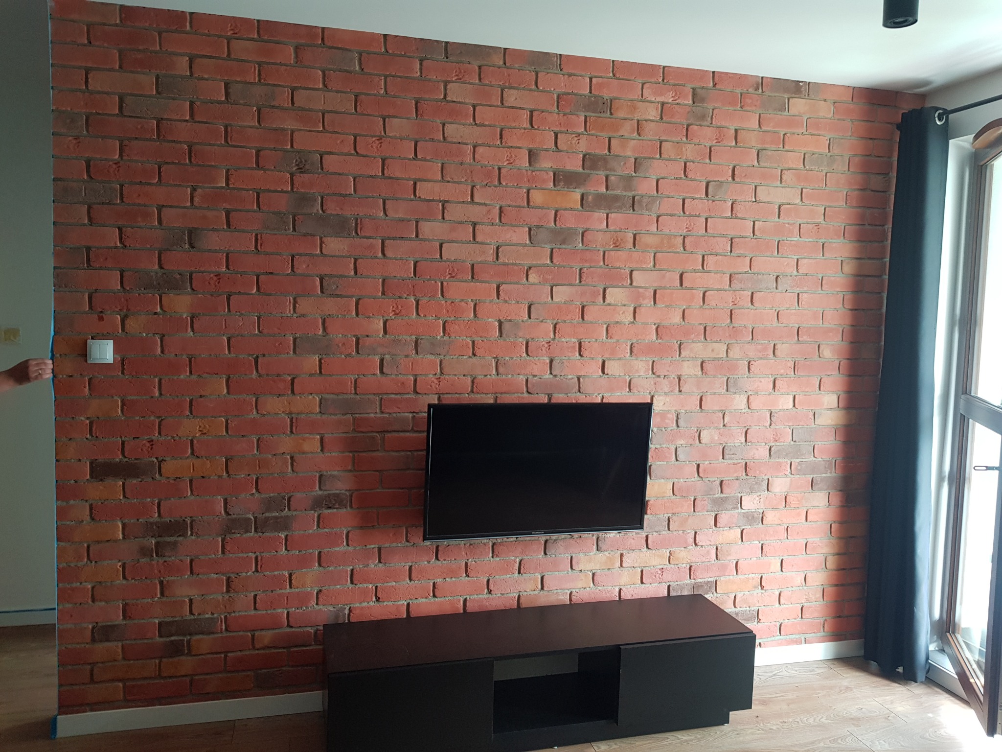 Ściana z czerwonej cegły w salonie z zawieszonym telewizorem i czarną szafką RTV, widoczne okno z zasłoną i fragment białej ściany z niebieską taśmą malarską.