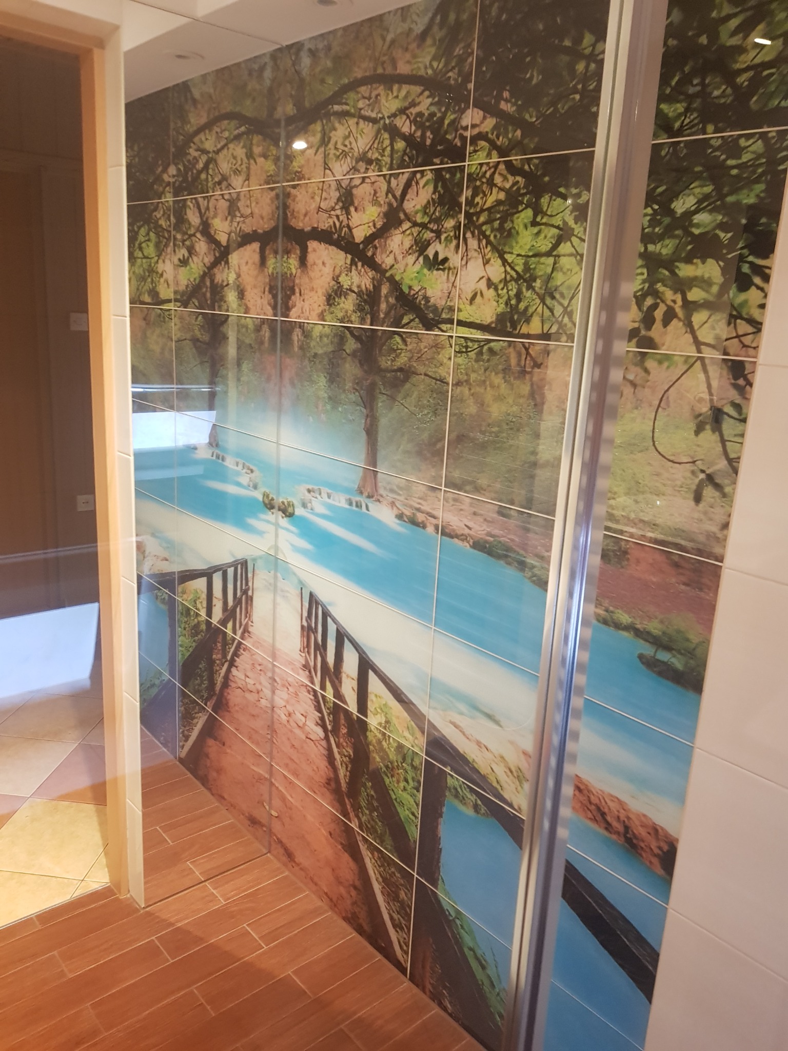 Ściana w łazience wyłożona płytkami z motywem krajobrazowym: drzewa i rzeka z mostem, dopasowane do drewnopodobnej podłogi