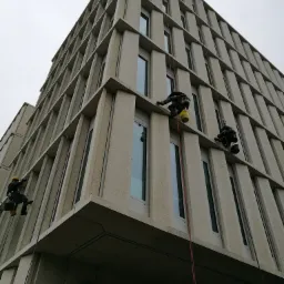 Trzech alpinistów na linach podczas prac na elewacji biurowca Bobrowiecka 8 w Warszawie, widok z dołu na budynek o regularnej, kanciastej architekturze.
