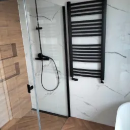 Nowoczesna kabina prysznicowa z czarnymi elementami, grzejnik drabinkowy w kolorze czarnym na ścianie z białych płytek imitujących marmur, podłoga wyłożona płytkami imitującymi drewno ułożonymi...